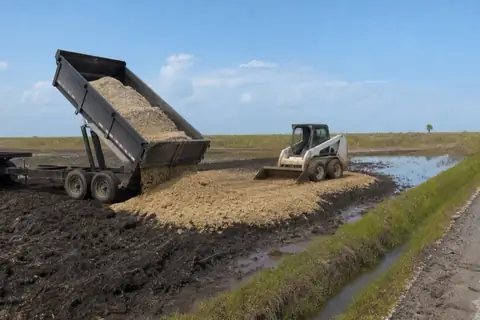 Trailer load of sandy fill being worked across the soggy end of a Pasadena lot by a compact loader