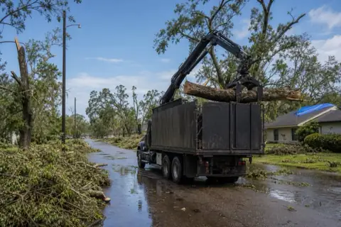 Grapple truck loading a snapped hackberry trunk from a storm-littered coastal yard
