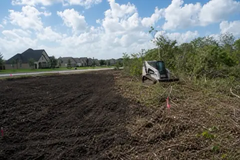 Quarter-acre subdivision lot half cleared to smooth dark clay with surveyor stakes and pink flagging